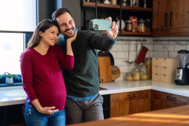 Sevimli bir adam hamile karısına sarılıyor, hamileliğinin son üç ayında selfie çekerek eğleniyor. Mutlu koca hamile kadını kucaklarken bebek bekliyor..