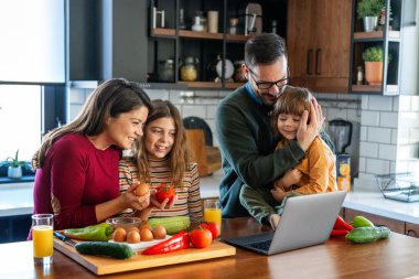 Evde dizüstü bilgisayar kullanan heyecanlı çocukları olan mutlu genç ebeveynler. Video konferansında sanal parti vermek, ekommerce ailesinin online alışverişini yapmak..