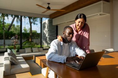Çok uluslu çift sabah rutininin keyfini çıkarıyorlar, gülümsüyorlar ve rahat ve modern bir ev ortamında dizüstü bilgisayar ve tablet kullanıyorlar.