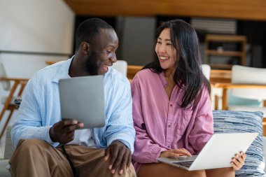 Çevrimiçi alışveriş veya ödeme için dijital dizüstü bilgisayar kullanan mutlu çift. Kadın kocasına dijital cihazdan bir şey alıyor. Orta Doğulu, Afro-Amerikalı çift dinleniyor..