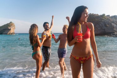 Genç yetişkinler, kumsalda parti yapan arkadaşlar soğuk içecekler ve büyük gülümsemelerle. Deniz kenarında kaygısız bir yaz kutlaması için mükemmel bir sahne.