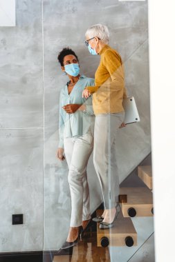 Teknoloji, yapay zeka, dijital yenilik ve salgın zamanlarında güvenli işyeri işbirliği için dizüstü bilgisayar kullanan maskeli kadın profesyoneller