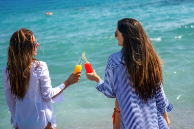 Two happy women toasting colorful beach drinks under the sun, enjoying tropical vibes and friendship moments by the sea