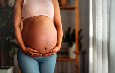 Mutlu bir yetişkin spor kıyafetleri içinde güzel hamile bir kadın oturma odasında dururken karnına sevgiyle bakıyor..