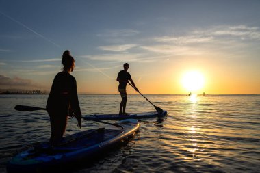 Sıcak günbatımı ışığında ayakta kürek çeken bir grup macera dolu aktif bir tatili yansıtıyor, açık hava refahı, doğada rahatlatıcı okyanus anları.