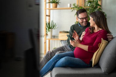 Hamilelik, teknoloji ve insan kavramı. Mutlu adam ve evde akıllı telefonları olan hamile karısı. Yeni mutlu aile.