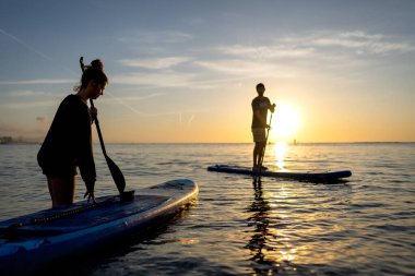 Sıcak günbatımı ışığında ayakta kürek çeken bir grup macera dolu aktif bir tatili yansıtıyor, açık hava refahı, doğada rahatlatıcı okyanus anları.