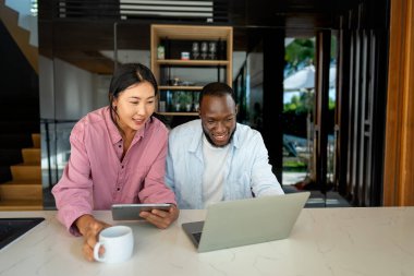 Genç ve çok ırklı çift evde bilgisayar ve dijital cihaz kullanırken eğleniyor. İnsanlar, teknoloji, online yaşam tarzı konsepti