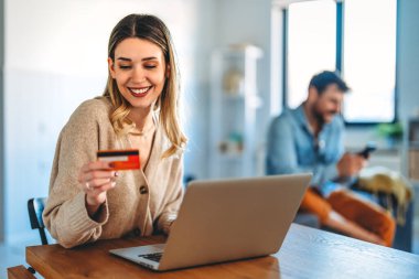 Gülümseyen bir kadın elinde bir bilgisayar ile evde bir kredi kartı ve alışveriş yapıyor, modern e-ticaret, dijital ödeme ve uygun online satın alma deneyimi temsil ediyor