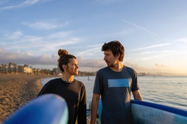 İki arkadaş gün batımında sahilde neşeli bir anı paylaşıyor, canlı sörf tahtaları tutarken aynı zamanda gülüyor ve okyanus güzelliğinin tadını çıkarıyor.