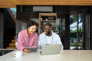Çok uluslu çift sabah rutininin keyfini çıkarıyorlar, gülümsüyorlar ve rahat ve modern bir ev ortamında dizüstü bilgisayar ve tablet kullanıyorlar.