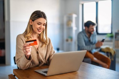Gülümseyen bir kadın elinde bir bilgisayar ile evde bir kredi kartı ve alışveriş yapıyor, modern e-ticaret, dijital ödeme ve uygun online satın alma deneyimi temsil ediyor