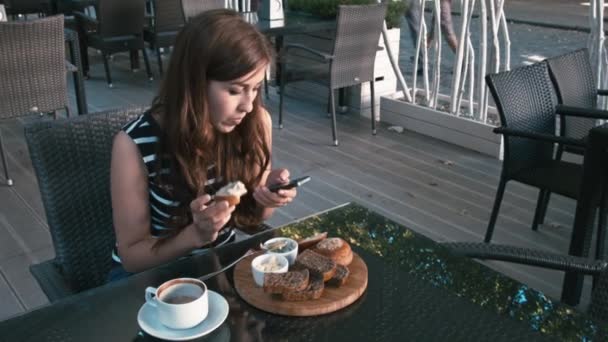 Mujer elegante mensajes de texto en el teléfono inteligente y comer un panecillo — Vídeo de stock
