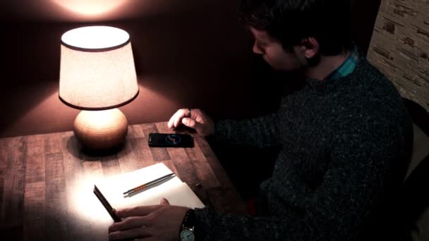 Der Typ am Schreibtisch mit dem Smartphone-Stift schreibt ins Notizbuch — Stockvideo