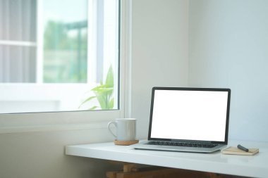 Simple workspace with white screen laptop, coffee cup and notebook on white table.	