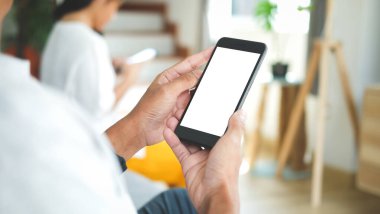 Boş ekranlı cep telefonu kullanan ve evdeki oturma odasındaki kanepede oturan genç bir adamın görüntüsü..