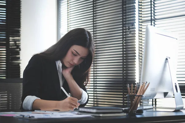 一位穿着黑色西服的快乐的女商人坐在现代化的办公室里 一边与电脑打交道 一边在笔记本上记笔记 — 图库照片