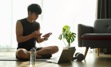 Evde egzersiz yaparken kulaklık takan ve akıllı telefon kullanan genç bir adam..