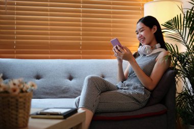 Akıllı telefonlu bir kadın kanepeye oturur ve evde mutlu bir şekilde telefonu kullanır..