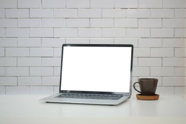 Boş ekranlı dizüstü bilgisayar ve tuğla duvarlı beyaz masada kahve fincanı..