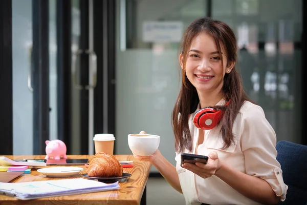 Genç Asyalı kadın kahve fincanı tutuyor ve cep telefonu kullanıyor..