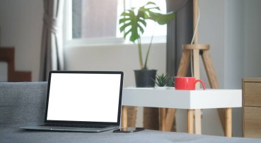 Boş ekranlı ve cep telefonlu açık bir dizüstü bilgisayarın görüntüsü modern oturma odasındaki kanepeye konuyor..