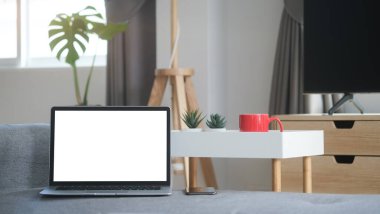 Boş ekranlı ve cep telefonlu açık bir dizüstü bilgisayarın görüntüsü modern oturma odasındaki kanepeye konuyor..