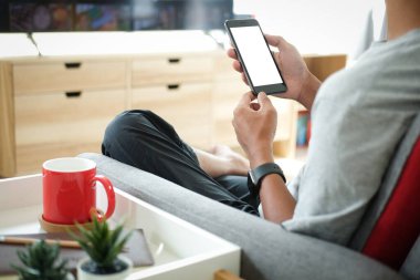 Elinde boş ekranla akıllı telefonu tutarken rahat koltukta dinlenen genç adamın kırpılmış görüntüsü..