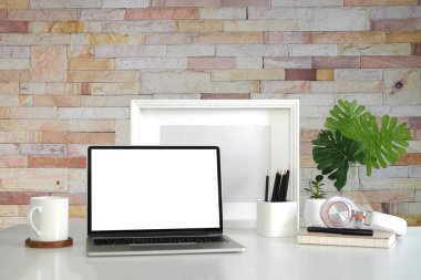 Laptop bilgisayar, kahve fincanı ve boş resim çerçevesi beyaz masada, tuğla duvarlı..