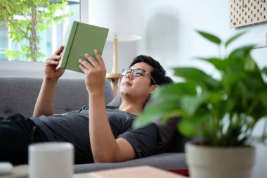 Huzurlu Asyalı adam rahat bir kanepeye uzanıp kitap okuyor..