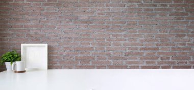 Boş poster çerçevesi, kahve fincanı ve tuğla duvarlı beyaz masa üzerine saksı bitkisi olan şık çalışma alanı.