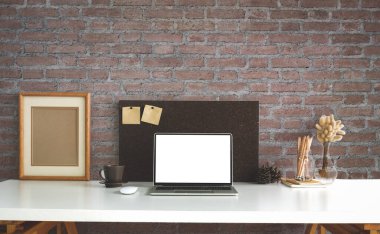 Öndeki bilgisayar dizüstü bilgisayarı, boş çerçeve ve beyaz masada tuğla duvarlı kahve fincanı..
