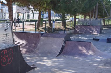 Çeşitli beton rampalar ve engellerle dolu boş bir kaykay parkının geniş bir görüntüsü. Bölge çam ağaçları ve metal bir çitle çevrili. Sabahları yumuşak gölgeler yerde uzanıyor..
