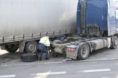 Yüksek görüş mesafesi olan bir kamyon şoförü mavi hidrolik kriko kullanarak hasarlı bir kamyonun lastiğini değiştirmek için yol kenarında çalışıyor..