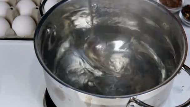 Ricetta per cucinare le uova in camicia. L'uovo crudo viene messo in acqua bollente in casseruola, dopo aver mescolato l'acqua con il cucchiaio. E lo fanno bollire. Primo piano. — Video Stock
