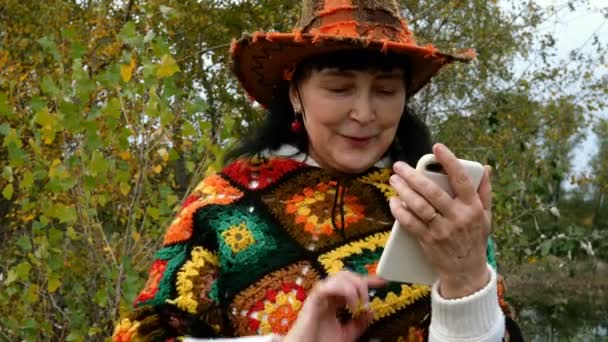カラフルな服のヴログでエレガントな成熟した大人の女性や彼女の顔の近くにそれを保持し、彼女のスマートフォン上のビデオ通話を行います。秋の公園や森で。閉鎖. — ストック動画