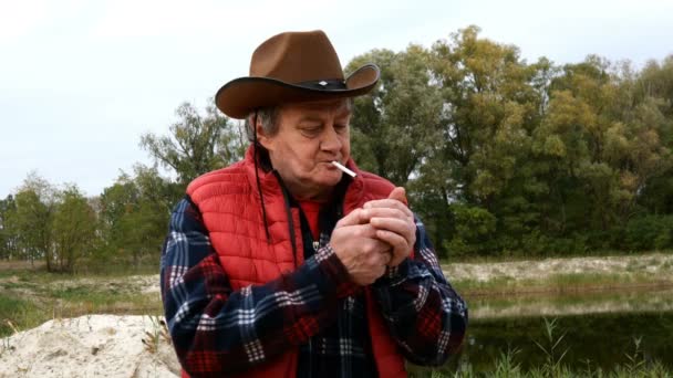 Volwassen volwassen man met cowboyhoed en -kleding, brandende sigaret van aansteker en inhaleert met plezier rook. Op het platteland of op de ranch. Middelmatig schot. — Stockvideo