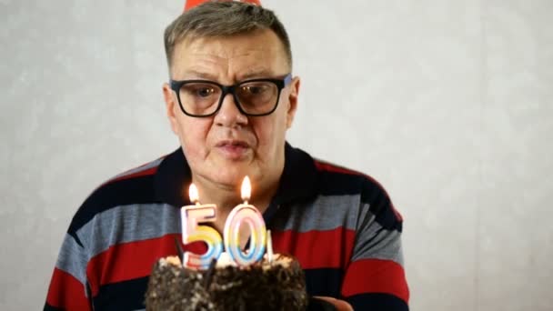 Joyful mature adult man wearing blue eyeglasses blows out number 50 candles on birthday cake. Make a wish. On light background. — Stock Video