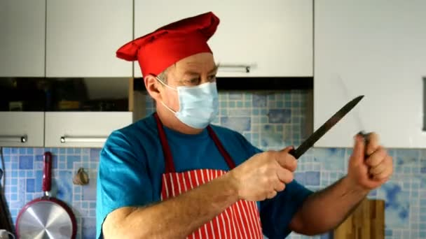Adulto mayor cocinero de etnia caucásica en chefs sombrero y mascarilla médica afilan cuchillo sobre cuchillo. — Vídeo de stock