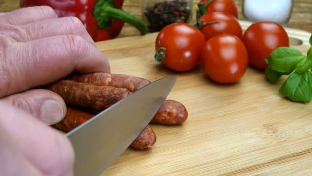 Las manos del chef cortan salchichas de caza en trozos con un cuchillo de cocina afilado en una tabla de cortar de madera para preparar deliciosa comida. Cocina casera. Primer plano. — Vídeo de stock