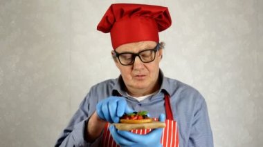 Kırmızı şef şapkalı, kıdemli yetişkin şef Kafkas kökenli, domatesli ve fesleğenli bruschetta 'nın tadını çıkarıyor, ev mutfağında. Yakın plan..