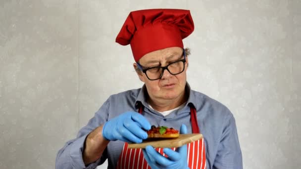 Senior chef adulto de etnia caucásica en sombrero de chef rojo, degustación con disfrutar de bruschetta con tomates y albahaca, en la cocina doméstica. Primer plano. — Vídeos de Stock