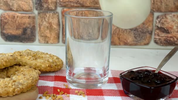 La leche fresca natural es vertida en el vaso de la jarra sobre la servilleta de la jaula. Cerca hay mermelada y galletas. Llenar el vaso con leche en el desayuno. Producto lácteo saludable concepto de alimentos. — Vídeo de stock