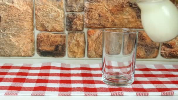 Fresh natural milk is poured into glass from pitcher against brick wall background on in cage napkin. Filling drinking glass with milk. Healthy dairy product food concept. Close-up. — Stock Video