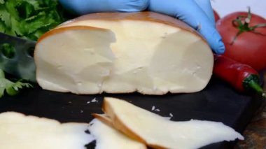 Büyük parça peyniri peynir bıçağıyla kesmek koyu ahşap kesim tahtasıyla, insan eliyle evcil mutfakta eldiven kullanmak. Domates, biber, dereotu, maydanoz. Yakın plan..