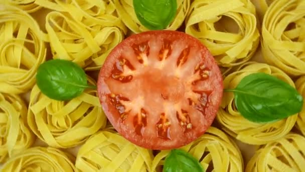 Veel droge tagliatelle pasta, versierd met basilicum bladeren en gesneden tomaat in het midden van de cirkel, draaien tegen de klok in. Bovenaanzicht opname. Close-up. — Stockvideo
