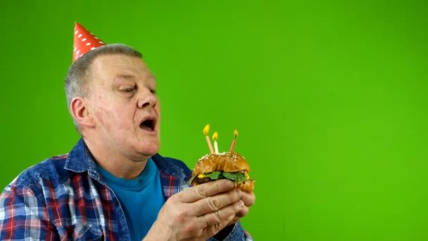 Взрослый мужчина белой национальности в праздничной кепке задувает свечи на гамбургере вместо праздничного торта и с удовольствием ест бургер. Хрома-ключ, зеленый экран. Крупный план. — стоковое видео