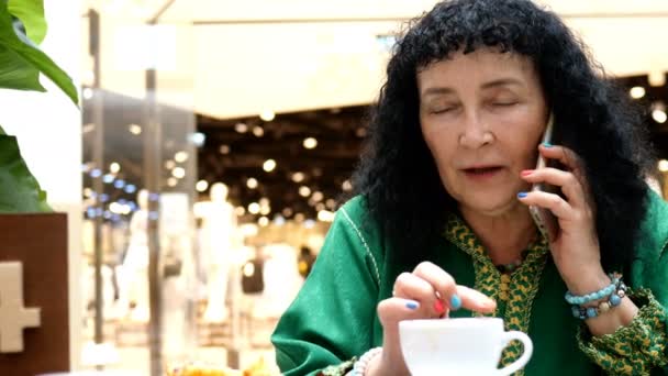 Portrait of brunette woman mature adult, caucasian ethnicity or mixed race, drinking coffee enjoying in public place and uses his smartphone. Focus on foreground. Close-up. — Stock Video