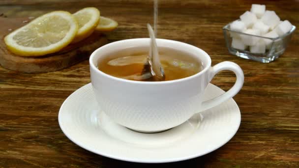 Bolsa de té elaborada en taza de cerámica blanca, sumergida en agua caliente hervida y dos piezas de azúcar refinada añadidas. Limón en rodajas se encuentra en la mesa de madera. Haciendo el desayuno inglés. Primer plano. — Vídeos de Stock