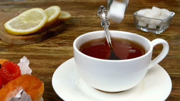 Två bitar raffinerat socker tillsätts i vit keramisk mugg med varmt te och rör om med tesked. Skivad citron och kaka ligger på träbordet. Göra engelsk frukost. Närbild. — Stockvideo